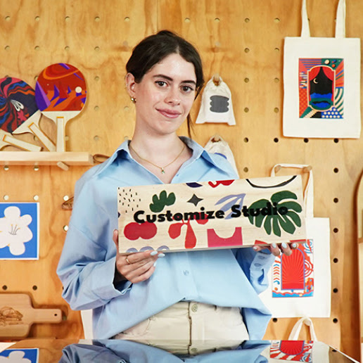 A woman showing a piece of wood with printed colourful shapes and the words 'Customize Studio' in black